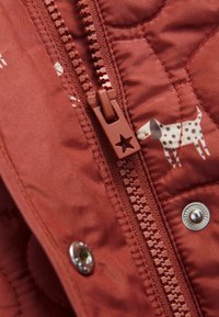Veste matelassée de couleur rouille avec un imprimé de chien. Présente une fermeture éclair marron avec un tirette en étoile et un détail de bouton-pression argenté.