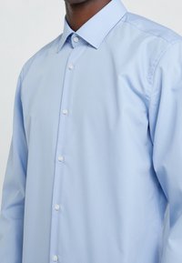 Lichtblauw langärmelig shirt van katoenen stof, met een klassieke kraag en een knoopsluiting aan de voorkant met parelachtig ogende knopen.
