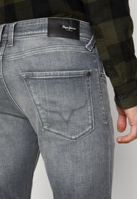 Grå denimjeans med en distinkt design på bakfickan, med en svart läderlogotyp vid midjan och en strukturerad yta.