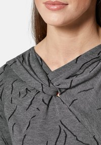Grijze top met een knoopdetail bij de halslijn en zwarte abstracte patronen. De stof lijkt zacht en heeft een gladde textuur.