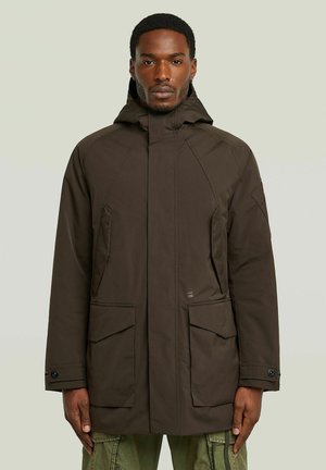 Homme de face portant une veste d’hiver à capuche marron foncé avec de grandes poches avant et un pantalon cargo vert.