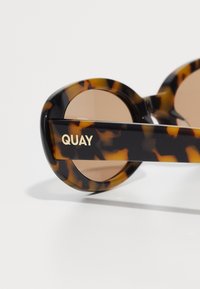 Iekrāsotu saulesbrilles ar apaļām rāmēm un brūnām lēcām. Uz sāniem ir zelta "QUAY" zīmols. Gluda plastmasas materiāls.
