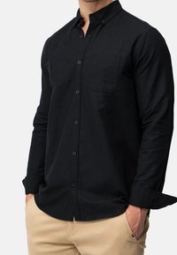 Chemise noire à manches longues avec boutons, poche poitrine, fabriquée en tissu doux, dotée d'un col et de poignets boutonnés. Associée à un pantalon beige.