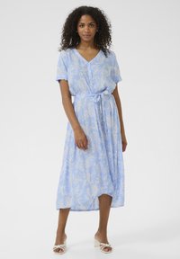 Abito maxi azzurro chiaro con maniche corte, scollatura a V, motivo floreale paisley, cintura da annodare in vita e tessuto morbido. Indossato con sandali a blocchi bianchi.