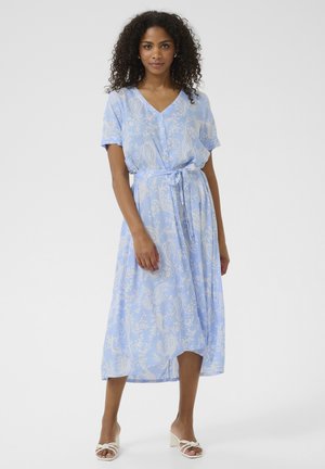 Light blue maxi dress with short sleeves, V-neck, floral paisley pattern, tie belt at waist, and smooth fabric. Worn with white block-heeled sandals.