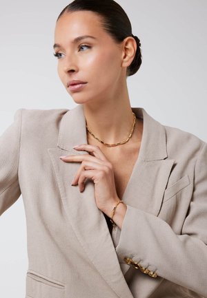 Goudkleurige sieraden hebben een delicate schakels ontwerp. Het onderwerp draagt een lichtbeige blazer van een gestructureerde stof met zichtbare knoopdetails.