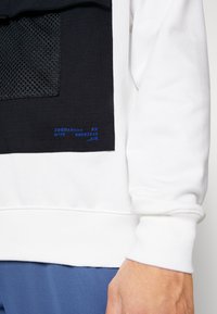 Hvid sweatshirt med en sort mesh-lomme, prydet med blå trykte tekst og accenter. Ribkanter ved ærmer og kant, blød tekstur, afslappet design.