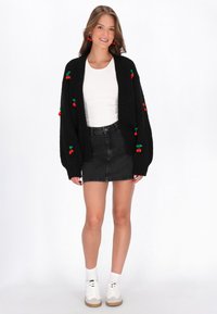 Juodas megztinis su raudonais pūkeliais, apsivilktas ant baltos bluzos ir juodų džinsinio audinio sijonų, derinamas su baltais sportbačiais. Kasdienis fitas ir tekstūruotas audinys.