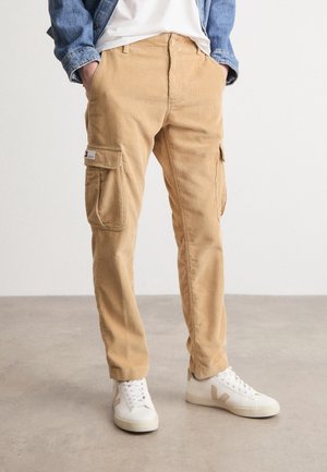 Persona con pantalones cargo de corduroy color beige, zapatillas blancas con logo beige, una camiseta blanca y una chaqueta de mezclilla azul con las manos en los bolsillos.
