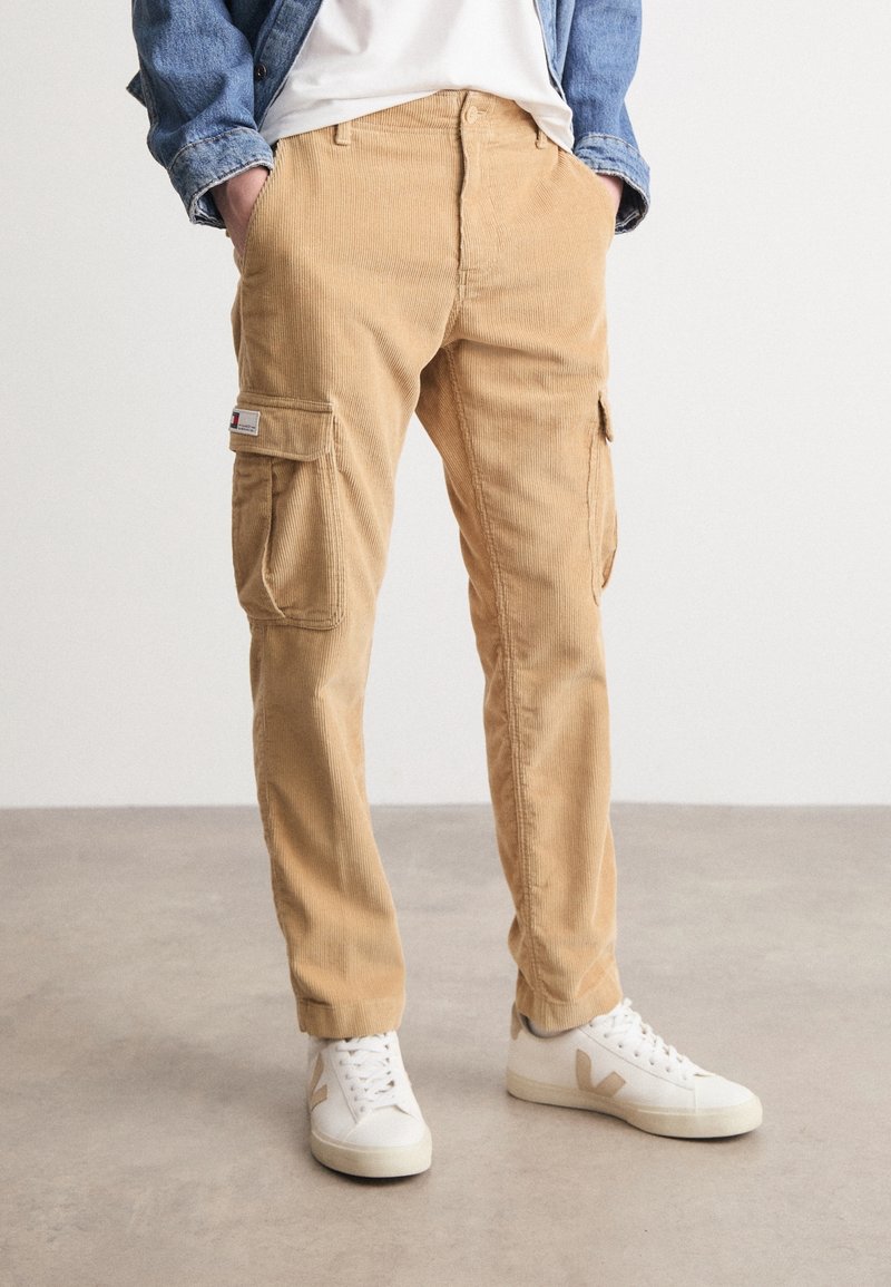 Henkilö, joka on pukeutunut vaaleanruskeisiin corduroy cargo-housuihin, valkoisiin lenkkareihin, joissa on beige logo, valkoiseen t-paitaaan ja siniseen farkkutakkiin, kädet taskuissa.