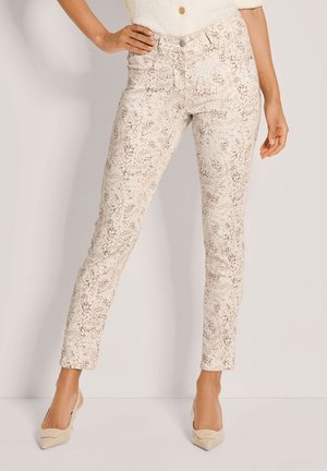 Frau trägt beige Slim-Fit-Hose mit hellbraunem Schlangenmuster und beige spitze High Heels, steht mit einer Hand in der Hüfte.