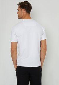 Camiseta blanca de manga corta con un acabado texturizado, que presenta un cuello redondeado y un pequeño acento rectangular en la parte superior de la espalda.