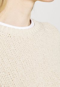 Crème-kleurige gebreide trui met een losse, gestructureerde ste stitch. Heeft een ronde hals en geribde manchetten, wat een zachte, comfortabele stof benadrukt.