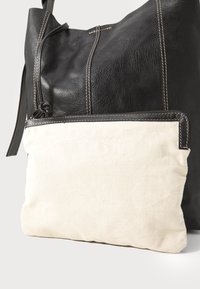 Svart lær totebag med teksturert overflate, med en kremfarget kanvaslomme foran med sydd detaljer. Justerbar stropp inkludert.