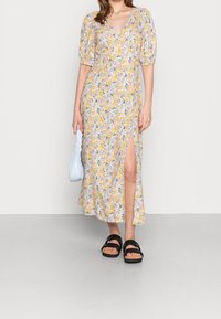 Robe midi florale avec un décolleté en V plongeant, des manches bouffantes, une base jaune et un imprimé de fleurs colorées. Présente une fente sur le côté, associée à des sandales noires et un sac bleu clair.