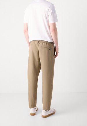 Mann trägt beige elastische Bundhose mit Reißverschlusstasche hinten und weißen Sneakers, steht vor schlichter weißer Wand.