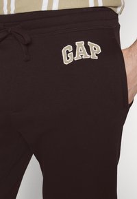 Dunkelbraune Sweatpants mit einem elastischenBund, ausgestattet mit einer Kordel und einem weißen gestickten "GAP"-Logo am linken Oberschenkel.