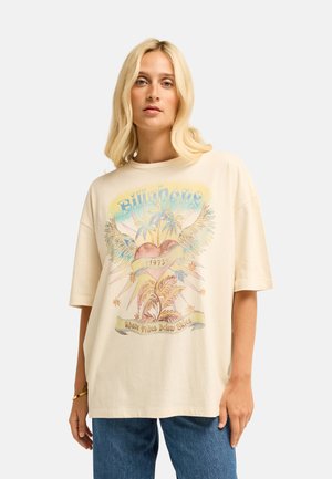 Blondine vrouw die een oversized beige T-shirt draagt met een kleurrijk vintage hart, palmbomen en tekst "Billabong" en "Above Tides Below Skies."