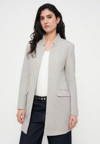 Ανοιχτό γκρι σακάκι από υφασμάτινο πλεκτό ύφασμα, με ψηλό γιακά, εμπρόσθιες τσέπες και σχεδίαση με ίσια γραμμή. Συνδυάζεται με λευκό τοπ και σκούρο τζιν.