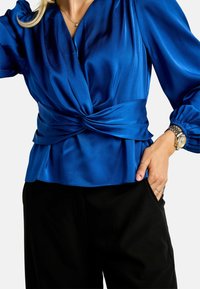 Modrá saténová blúzka s uzlom vpredu, dlhé nafúknuté rukávy a hlboký výstrih. Kombinovaná s čiernymi nohavicami.