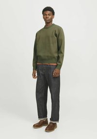 Suéter verde oliva con cuello acanalado, combinado con pantalones vaqueros azul oscuro y zapatos marrones con cordones. El conjunto tiene un ajuste relajado y puños enrollados.