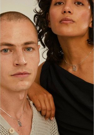 Silver kedja halsband med en geometrisk berlock. Närbild av två modeller, en i en strukturerad, ljus tröja och den andra i en mörk topp.