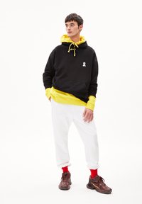 Felpa nera con accenti gialli e cordino, indossata sopra un felpa gialla. Pantaloni jogger bianchi e sneaker marroni con calzettoni rossi completano il look.