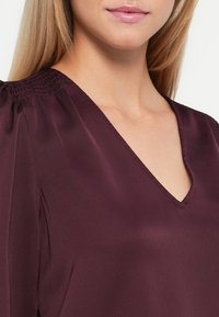 Bluză din mătase burgundy cu decolteu în V, având mâneci bufante și o textură subtilă la cusăturile umerilor. Material neted și fluid.