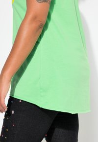 Green cotton t-shirt with a relaxed fit, showing a side view. Paired with black jeans featuring colorful decorative accents.
