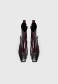 Une paire de bottines en cuir marron foncé, avec des orteils pointus et carrés, et des fermetures éclair latérales, vues d'en haut sur un fond blanc.
