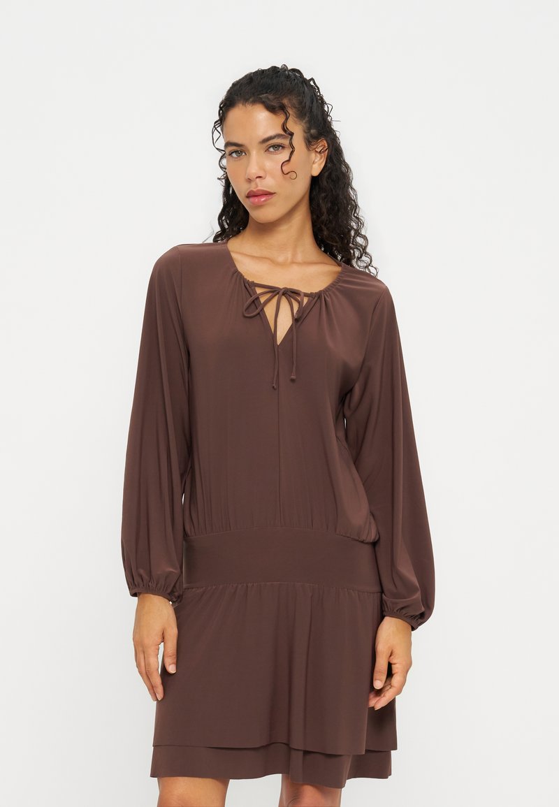 Femme aux cheveux bouclés et foncés portant une robe marron à manches longues avec un col à nouer, debout devant un fond clair uni.