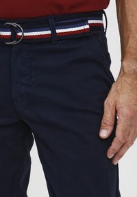 Pantaloni blu navy con una cintura intrecciata caratterizzata da strisce rosse, bianche e blu e una fibbia a D in metallo, che mostrano dettagli del tessuto testurizzato.