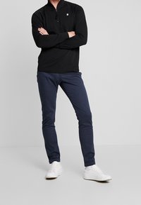 Svart långärmad polo med kvartdragkedja, marinblå slim-fit byxor och vita sneakers. Polon har en diskret logo nära kragen.