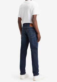 Jeans de denim oscuro con corte recto, bolsillos traseros con un patrón de costura, combinados con una camiseta blanca sencilla y zapatillas blancas.