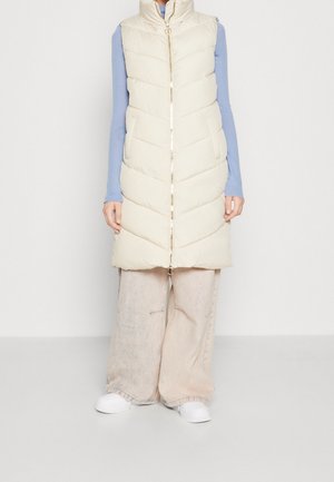Kvinde iført en cremefarvet quiltet, ærmeløs dunvest over en blå langærmet top, beige vidde bukser og hvide sneakers, stående.