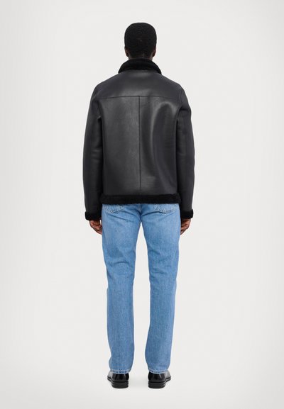 Veste en cuir noir avec un col en fourrure, présentant une coupe droite, un tissu texturé et des manches longues. Associée à un jean bleu clair.