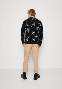 Juodas pūkuotas megztinis su baltu šakotų raštu, derinamas su smėlio spalvos sukrautais chinos kelnais ir juodais bateliais, matomas iš nugaros.