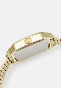 Montre-bracelet dorée avec un boîtier rectangulaire, une couronne hexagonale et un bracelet en métal. Le cadran est argenté avec un minimum de détails.