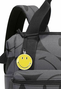 Mochila gris con un acabado texturizado, que presenta un llavero de cara sonriente amarilla adjunto. Se pueden ver correas duraderas y un compartimento con cremallera.