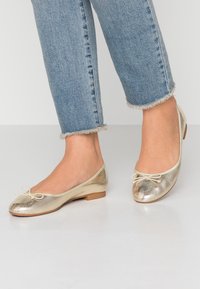 Guldmetalliska balettflat-former med rund tå, platt häl och en dekorativ rosett. Matchas med nedklippta ljusa denimjeans med fransad fåll.