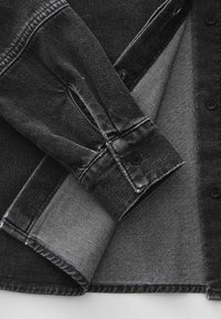 Schwarze Jeansjacke mit umgeschlagenem Ärmel, der die geknöpfte Manschette und die Innentextur gegen einen flachen weißen Hintergrund zeigt.