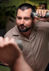 Electric hair clipper in hand, grey body, adjustable guard. Man with short, dark hair and light brown button-up shirt, focusing on grooming.