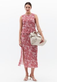 Vestido sin mangas con estampado floral en rosa y blanco, con un diseño de peplum y una abertura lateral. La modelo sostiene un bolso beige.