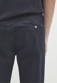 Pantaloni blu scuro con una texture liscia, due tasche posteriori, un dettaglio con bottone argentato e una piccola etichetta del marchio sul lato.