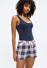 Γυναίκα με σγουρά μαλλιά που φοράει μπλε τοπ χωρίς μανίκια και μπλε-ροζ σκουρόχρωμα καρό σορτς με κορδόνι, στέκεται μπροστά σε έναν απλό λευκό φόντο.