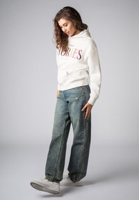 Witte hoodie met "STORIES" print in roze en paars, gecombineerd met losse blauwe denim jeans en witte sneakers. Casual, relaxed fit.