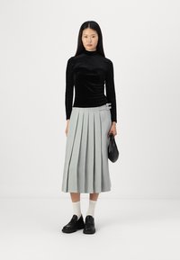 Haut long à manches noires en velours avec des épaules texturées, associé à une jupe midi plissée grise avec un détail de boucle en argent. Sac noir et chaussures.