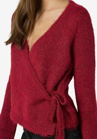 Kvinna klädd i en strukturerad djup röd insnörad kofta som är knuten i midjan med en rosett, tillsammans med svarta byxor, mot en neutral bakgrund.