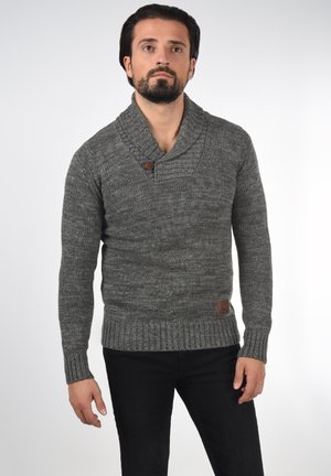 Homme aux cheveux foncés et à la barbe, portant un pull en laine gris à col châle avec bouton, et un pantalon noir, debout devant un fond uni.