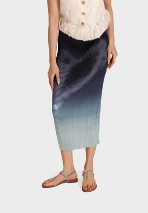 Vrouw die een geplooide midi-rok met een verloop van donker marineblauw naar lichtblauw draagt, gecombineerd met een witte blouse met ruches en zilveren platte sandalen.